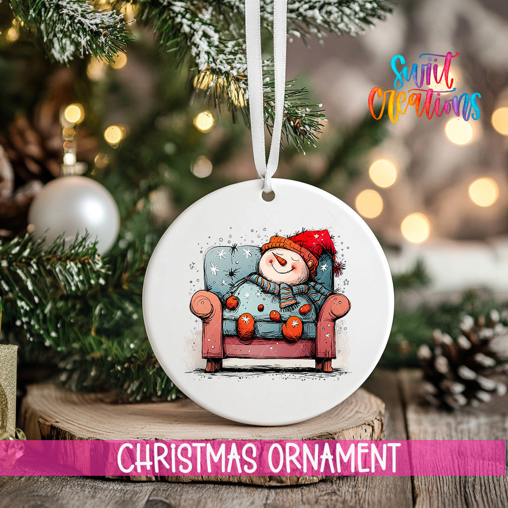 A white Christmas ornament with a snowman sitting in a chair, surrounded by snow and twinkling lights, is displayed on a wooden surface.