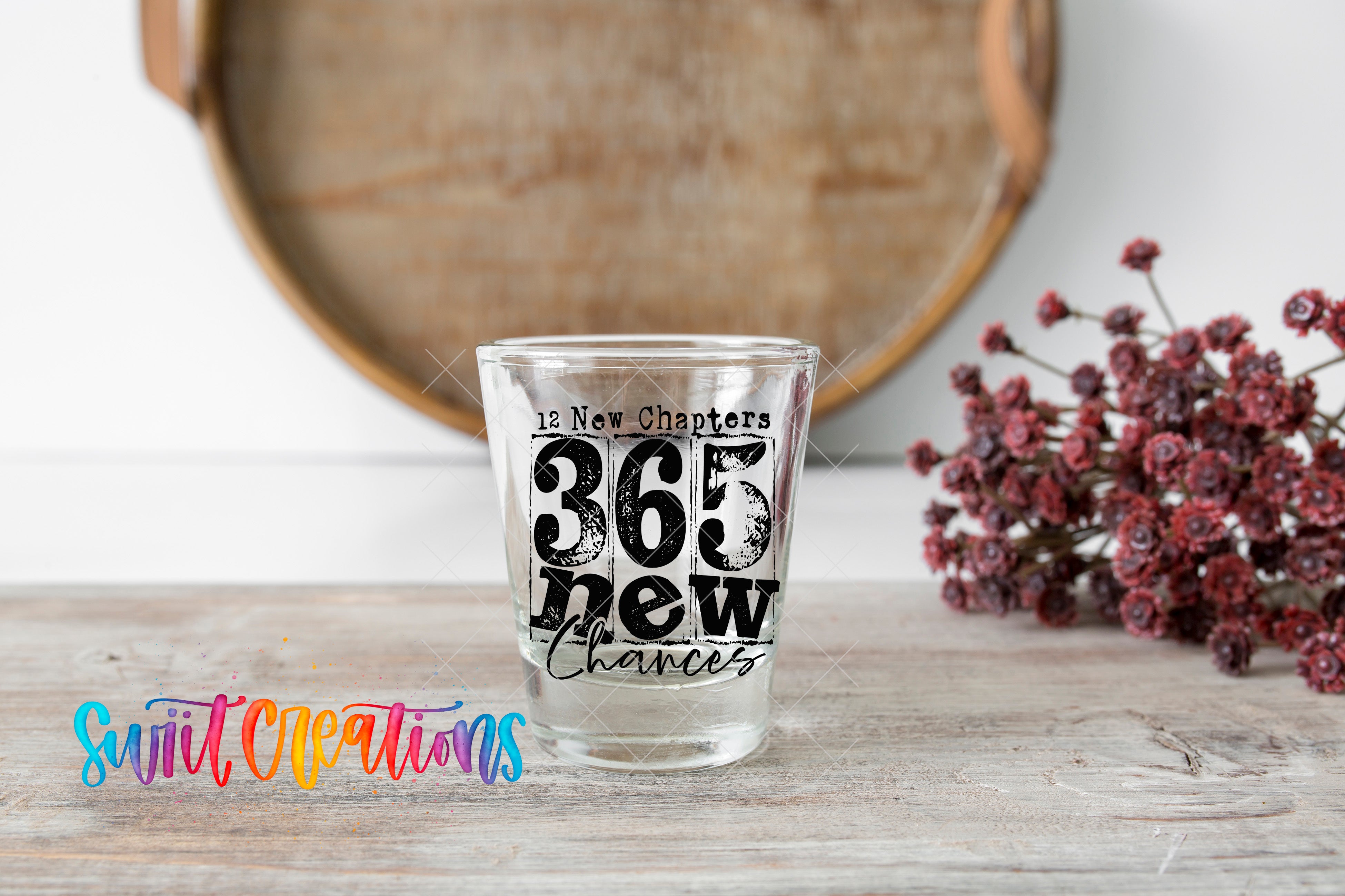 A shot glass with the text "12 New Chapters 365 New Chances" printed on it, placed on a wooden surface next to a bunch of red flowers.