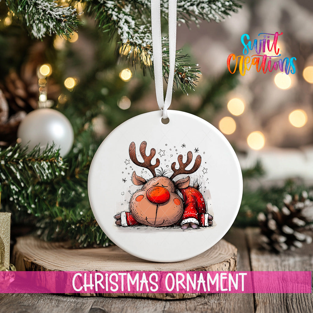 A white Christmas ornament with a cartoon reindeer design hangs on a tree branch, surrounded by pine cones and other holiday decorations.