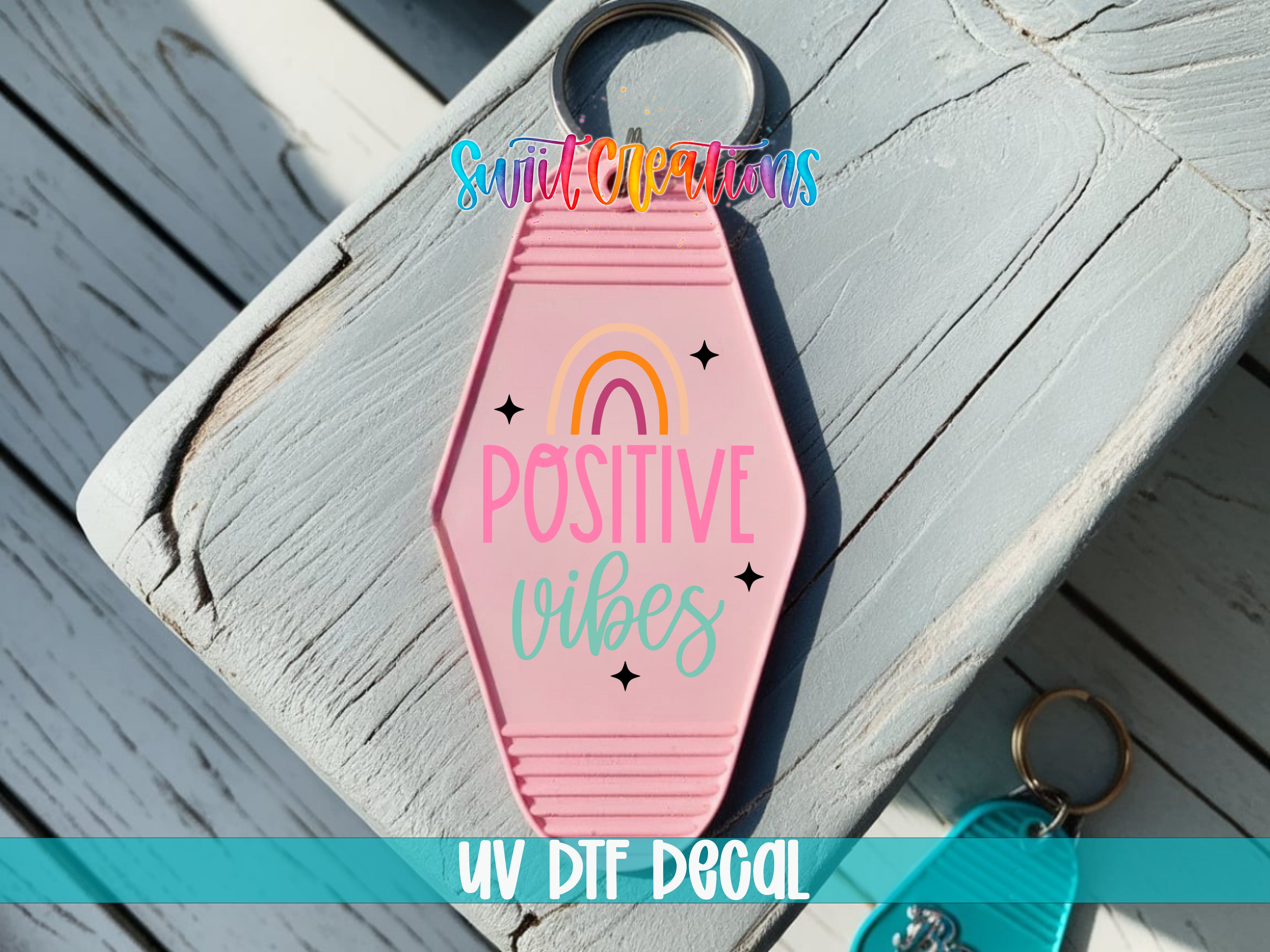 A pink keychain with the text "POSITIVE VIBES" printed on it, placed on a wooden surface.