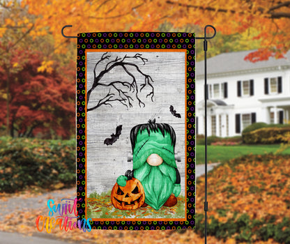 A Halloween-themed garden flag featuring a green gnome holding a jack-o-lantern, with a spooky tree and bats in the background.