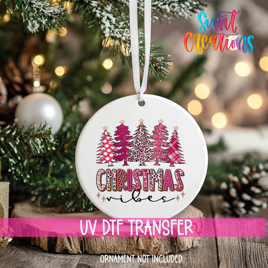 A white Christmas ornament with a pink and white tree design hangs on a tree branch, surrounded by other Christmas decorations.