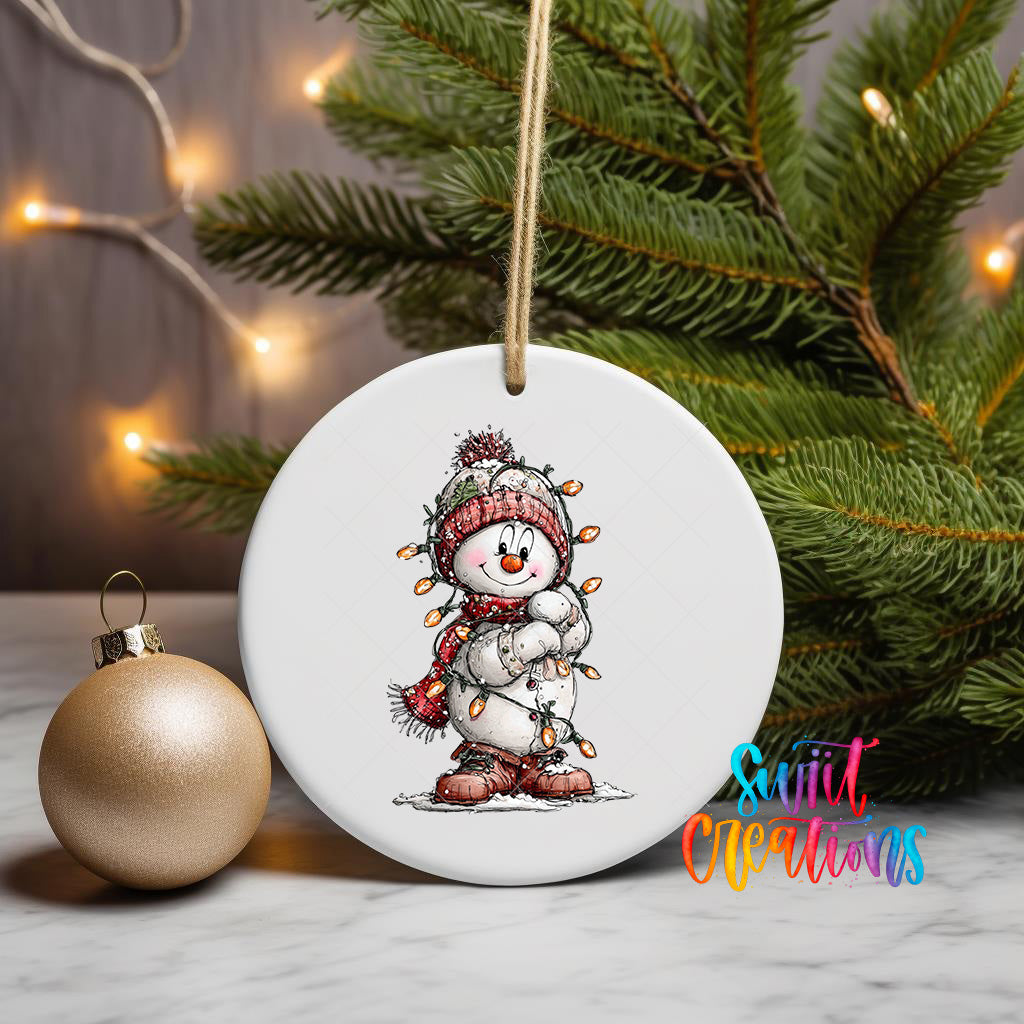 A white ceramic ornament featuring a smiling snowman is displayed on a table, with a gold ornament and a Christmas tree in the background.