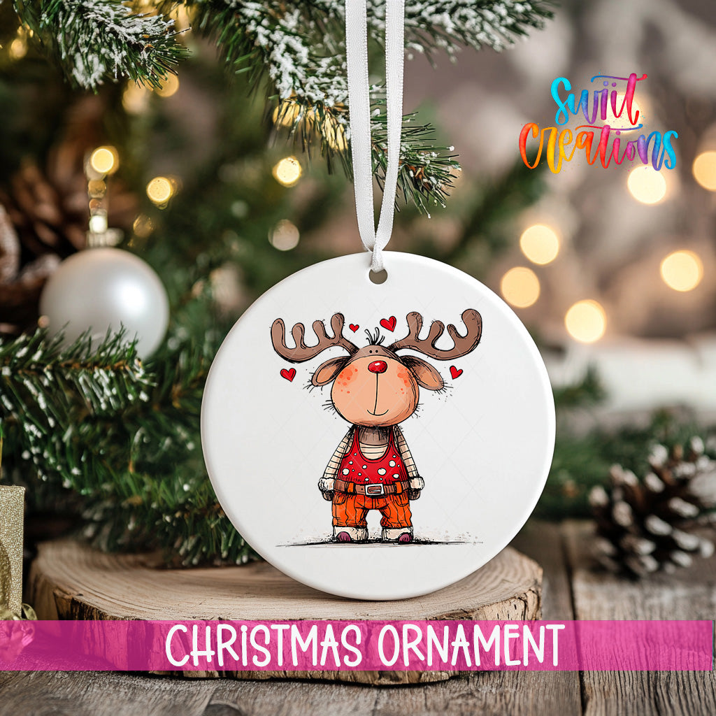 A white Christmas ornament with a cartoon reindeer and hearts hanging from a ribbon, placed on a wooden surface with a blurred background of a decorated Christmas tree.