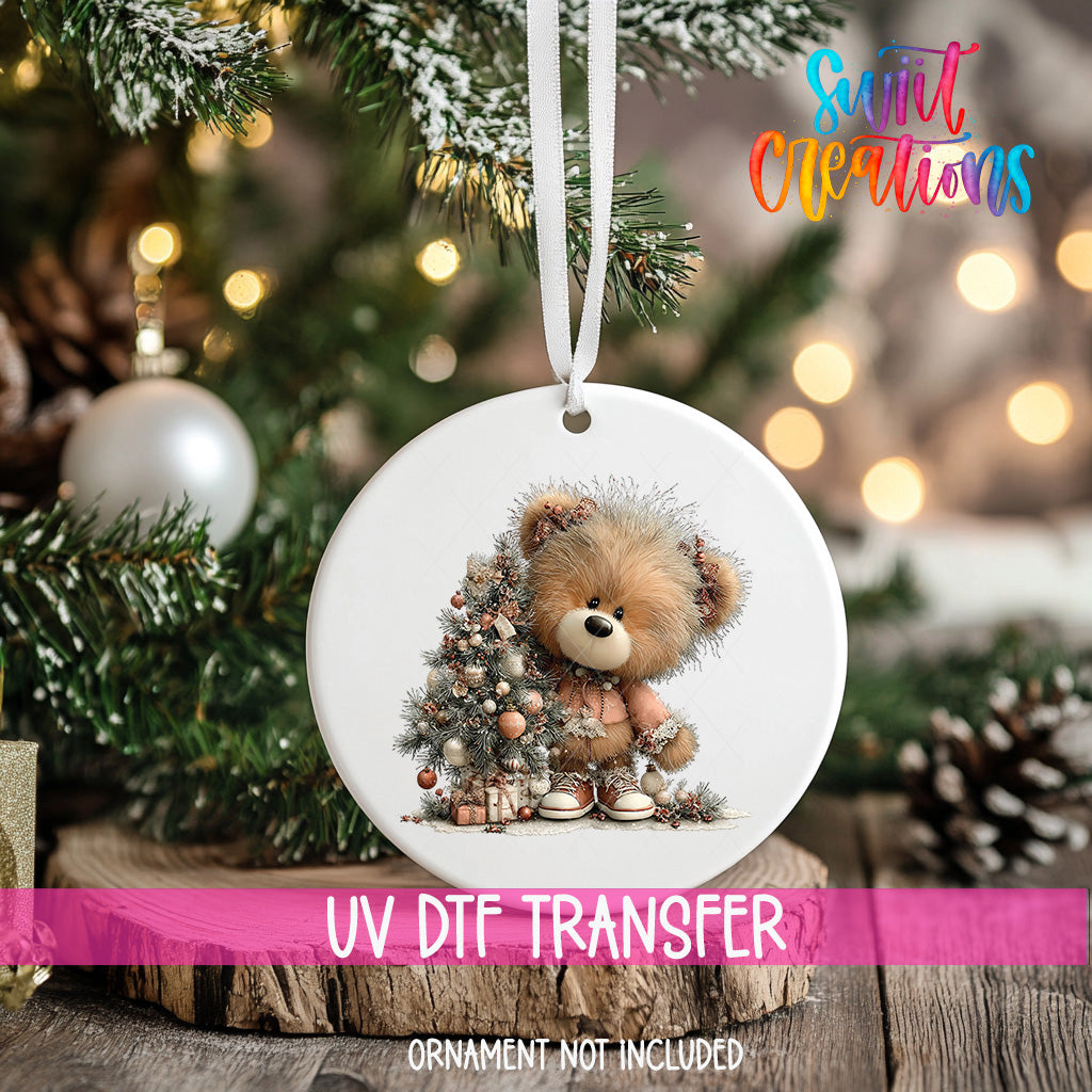 A white ornament with a teddy bear image is hanging on a Christmas tree, surrounded by pine branches and other holiday decorations.