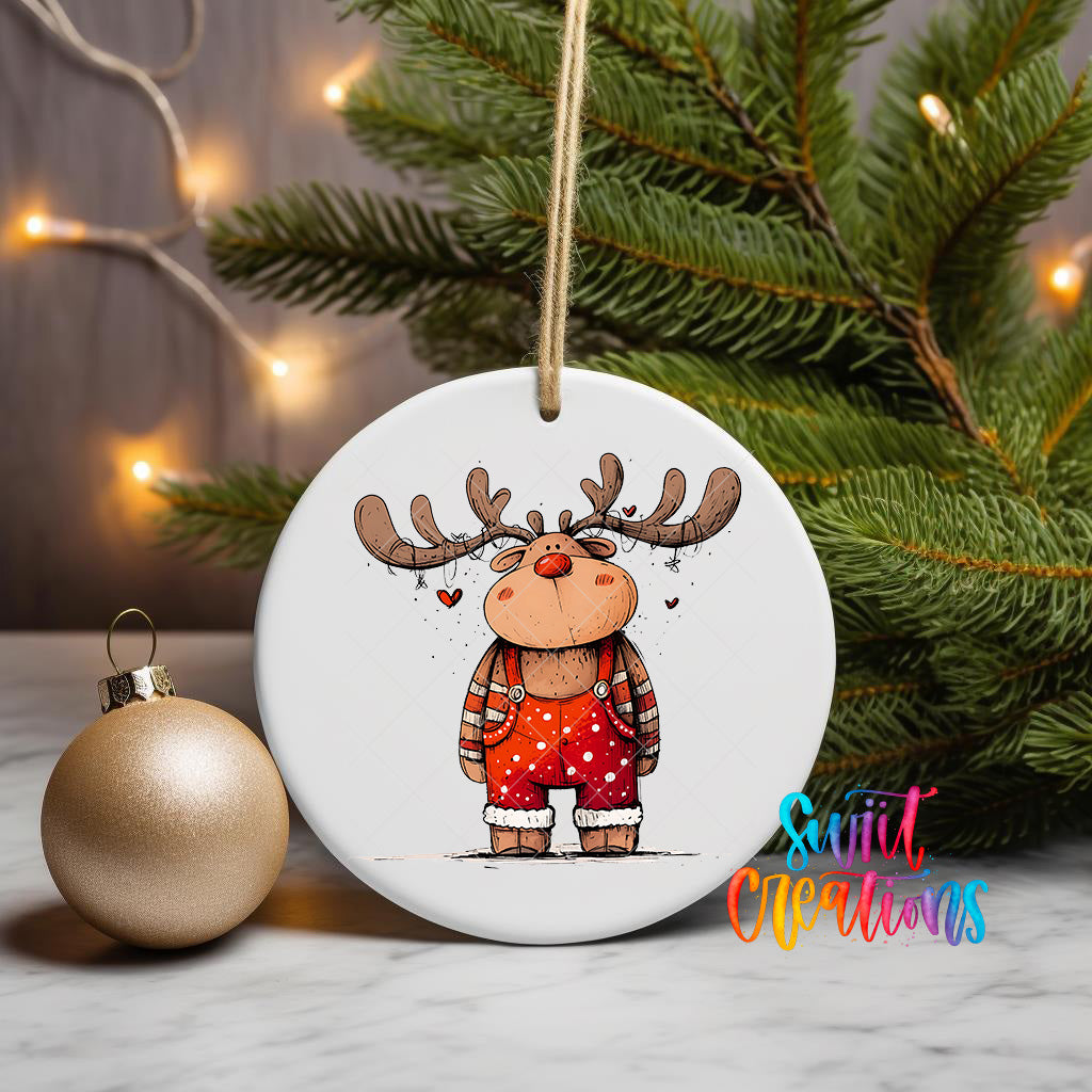 A white ceramic ornament featuring a cartoon reindeer wearing red and white polka dot pants, with a heart-shaped decoration hanging from a string. The ornament is placed next to a gold ornament and a Christmas tree, creating a festive holiday scene.