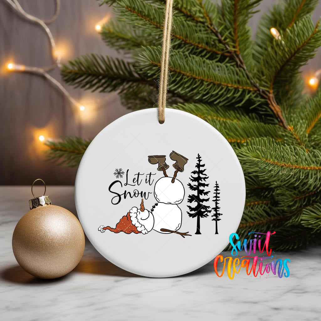 A white ceramic ornament with a snowman and pine trees, along with a gold ornament, is displayed on a table with Christmas lights in the background.