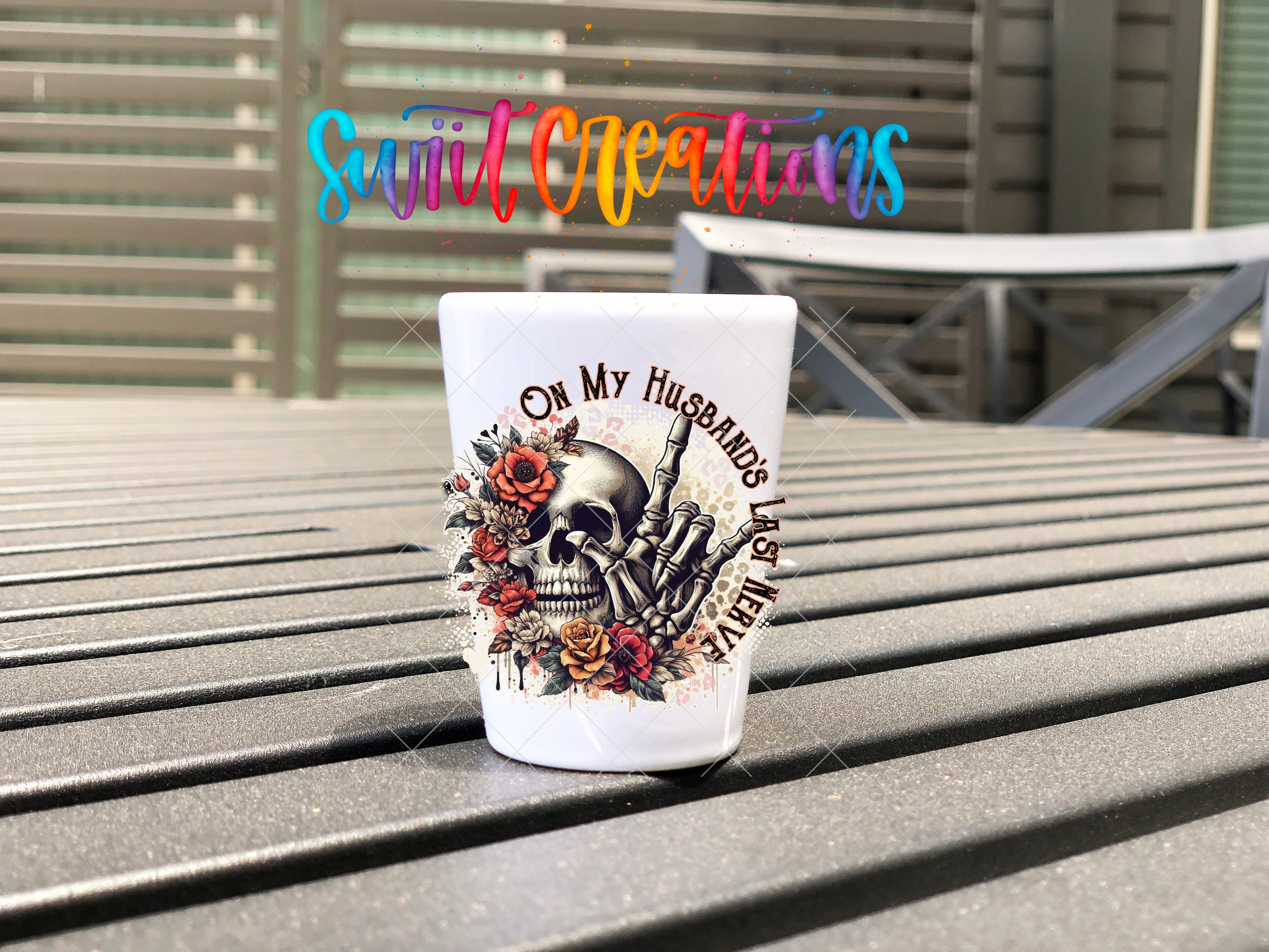 A white shot glass with a skull design and floral elements is placed on a wooden surface.