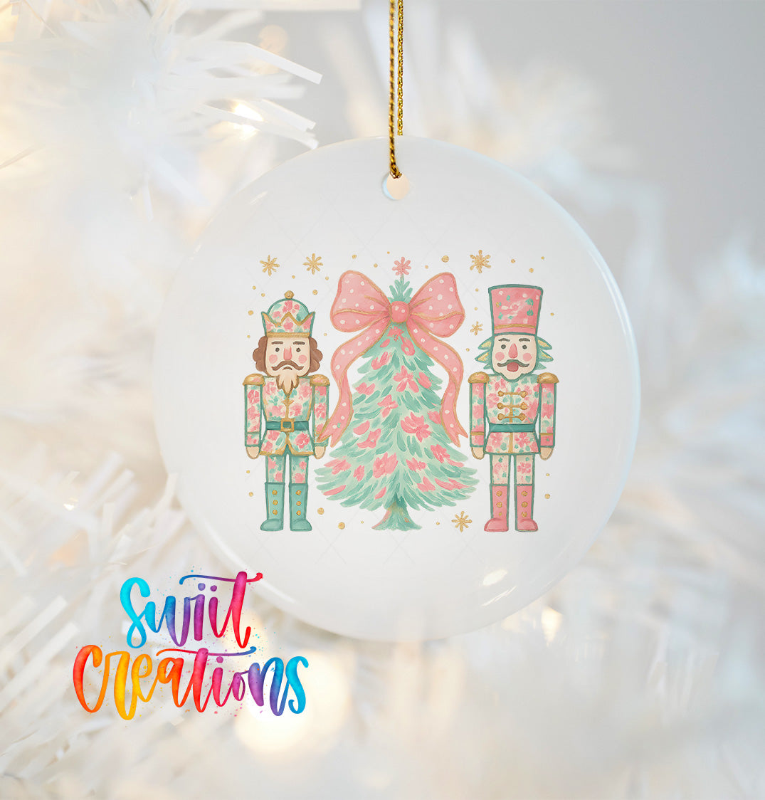 A white ornament with a Christmas tree and two nutcracker characters, one wearing a crown and the other a hat, hanging on a string.