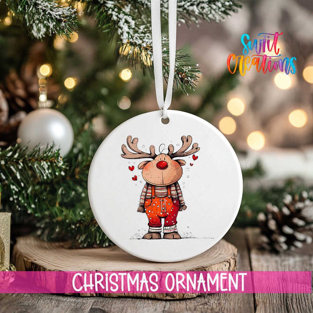 A white Christmas ornament featuring a cartoon reindeer with antlers and a red nose, hanging on a tree branch.