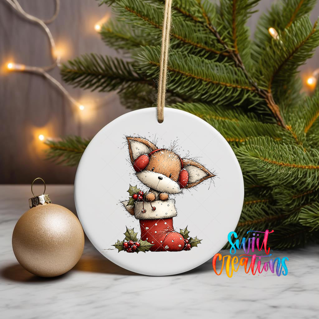 A white ceramic ornament featuring a cartoon image of a fox wearing a red stocking, sitting on a branch with holly leaves, is displayed on a table next to a gold ornament and a Christmas tree.