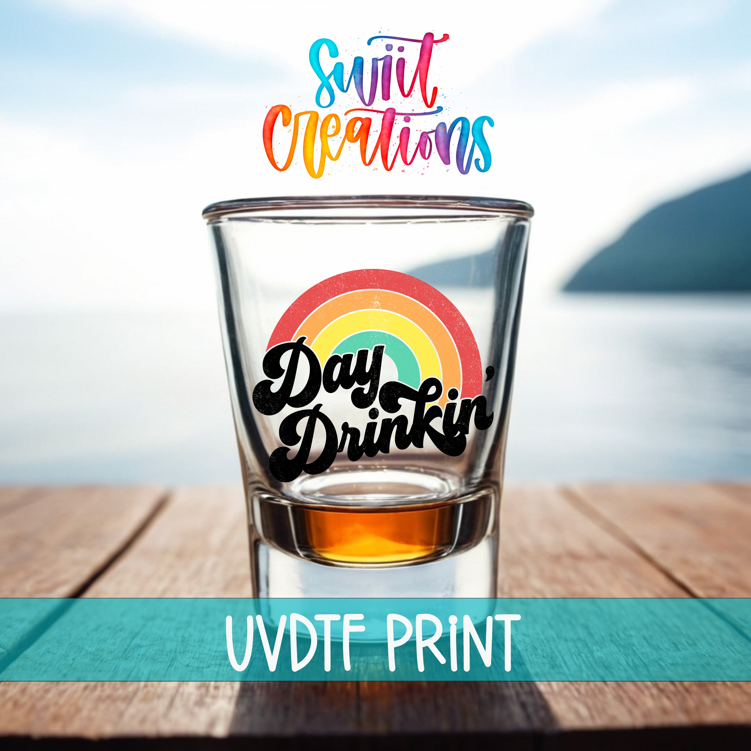 A shot glass with a rainbow graphic and the text "Day Drinking" printed on it, placed on a wooden surface with a body of water in the background.