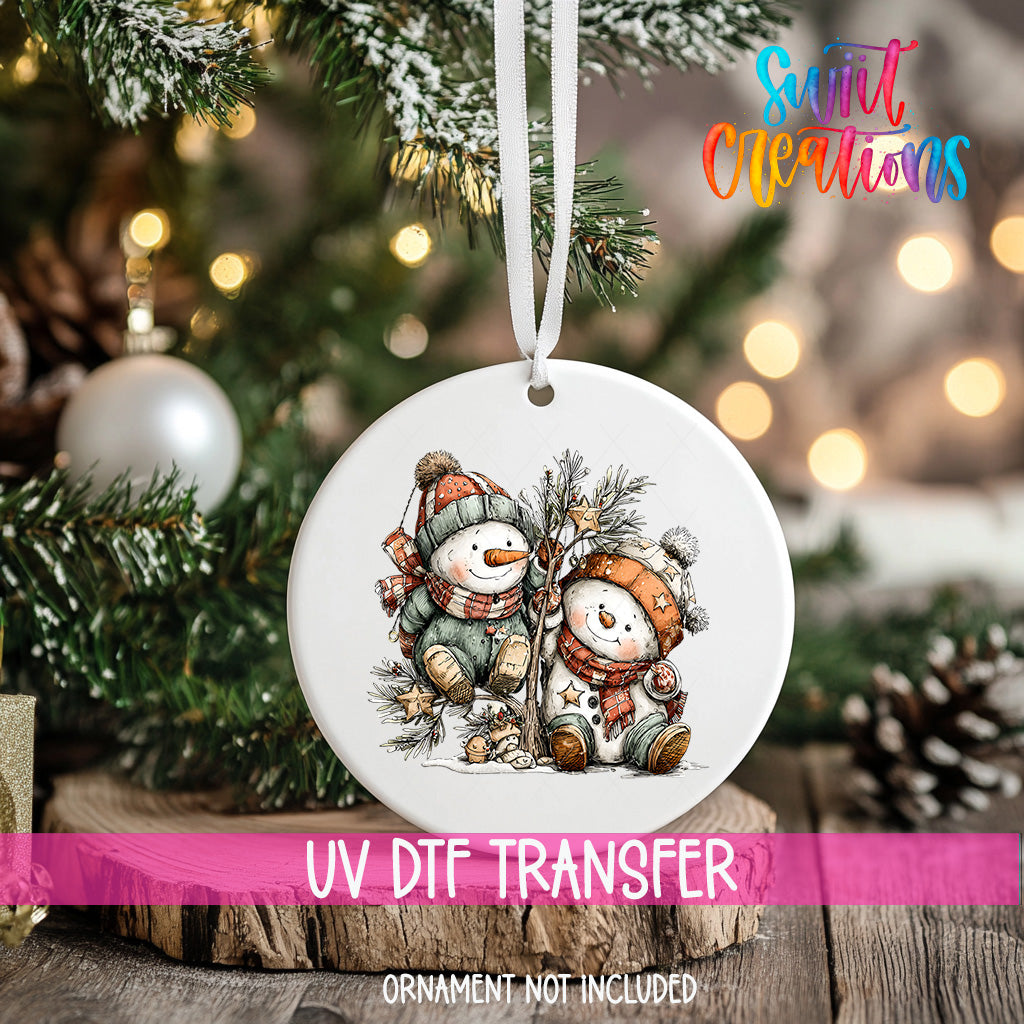 A white ornament with a snowman and pine tree design hangs on a Christmas tree, with a pink ribbon around it.