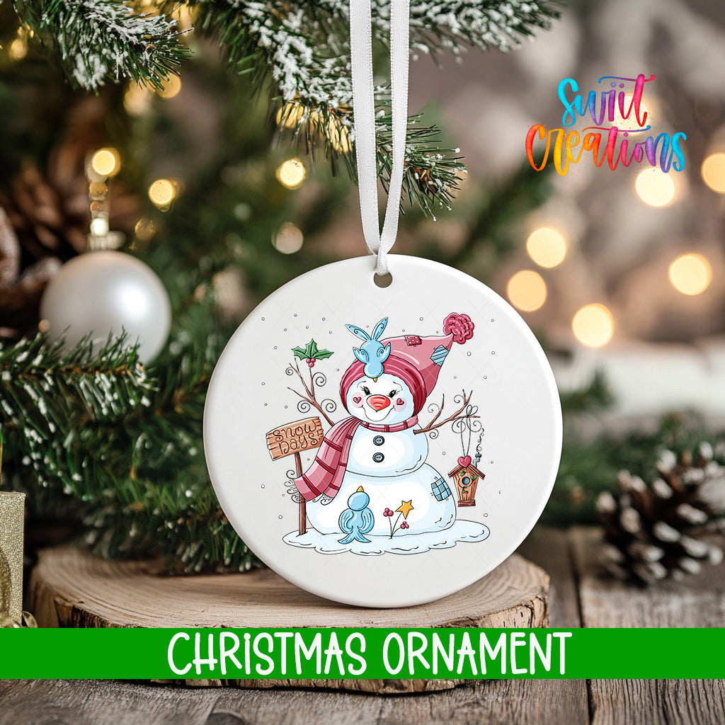 A white Christmas ornament featuring a snowman and other winter-themed elements hangs from a tree branch.