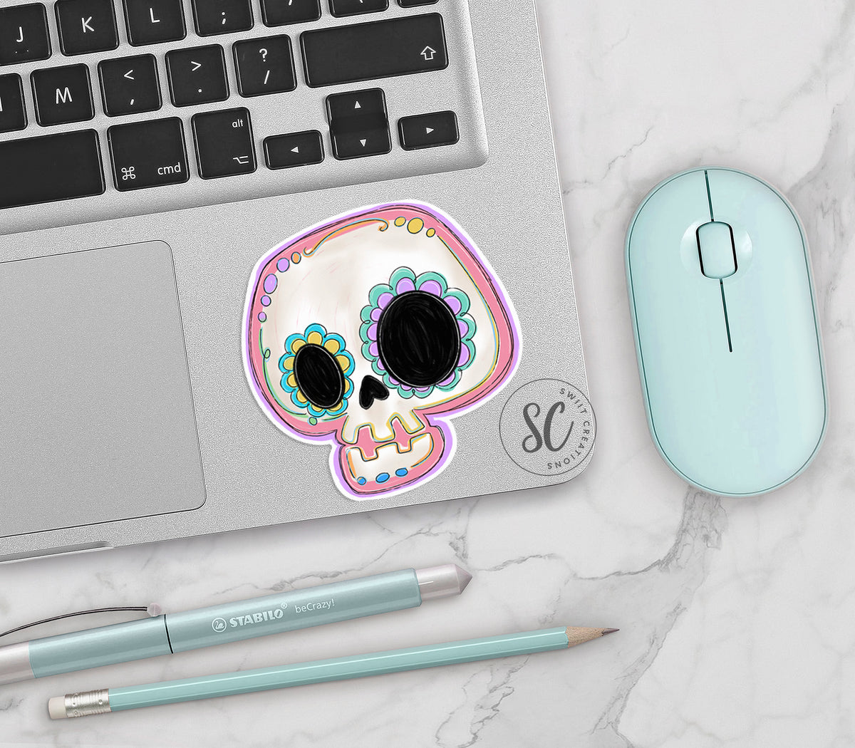 Pastel Sugar Skull vinyl sticker on a silver laptop next to a mint mouse and pens