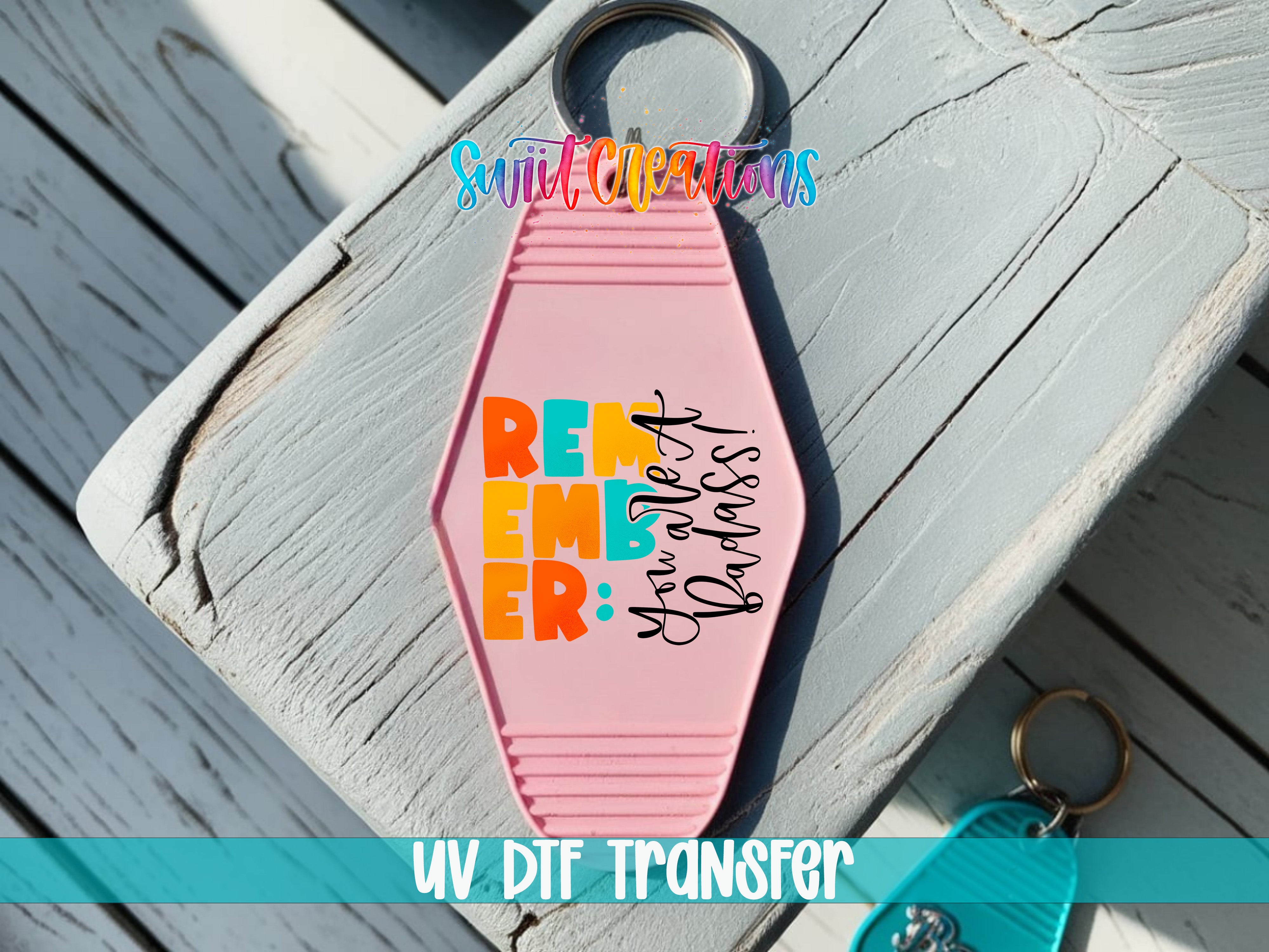 A pink keychain with the text "REMEMBER" and "You're Badass" printed on it, placed on a wooden surface.