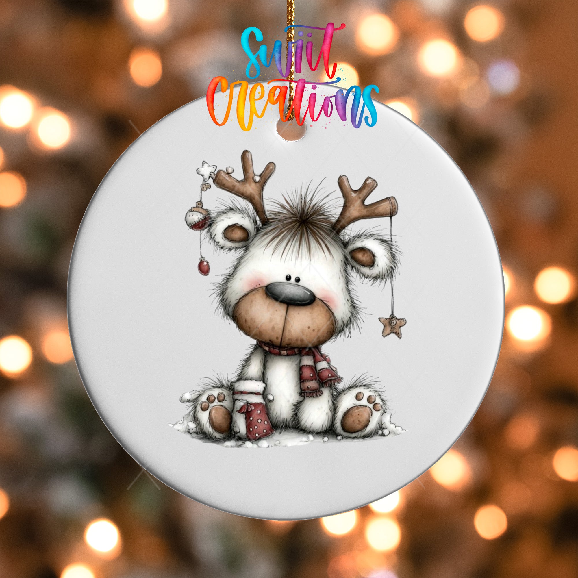 A white ornament with a cartoon reindeer and Christmas decorations, including a star and a snowflake, hanging against a blurred background with twinkling lights.
