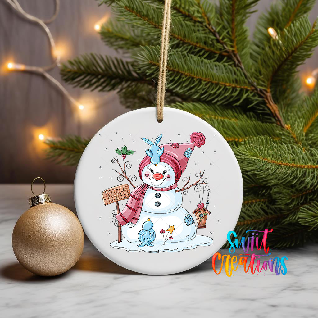 A white ceramic ornament featuring a snowman with a sign that says "Silly Creations" is displayed on a table next to a gold ornament and a Christmas tree.