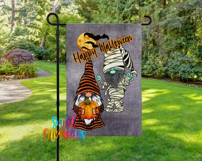 A Halloween-themed garden flag featuring two gnomes, one dressed as a mummy and the other as a gnome, holding pumpkins and standing on a grassy lawn.