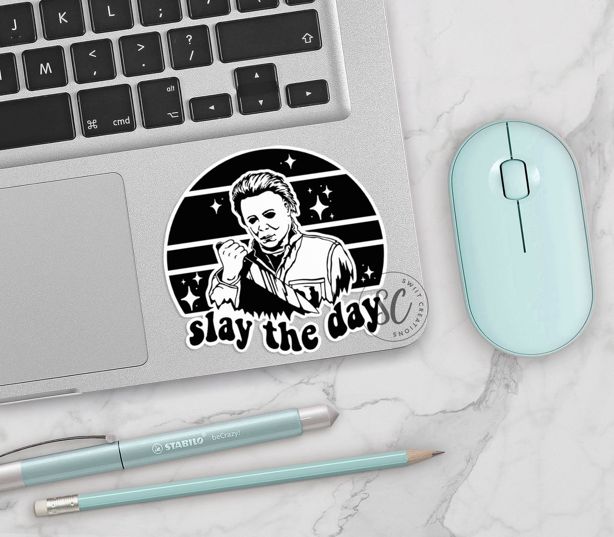 Slay the Day vinyl sticker on a silver laptop next to a mint mouse and pens