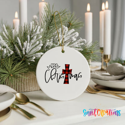 A white Christmas ornament with a red and black plaid cross design hangs on a table, surrounded by a festive arrangement of pine branches and candles.