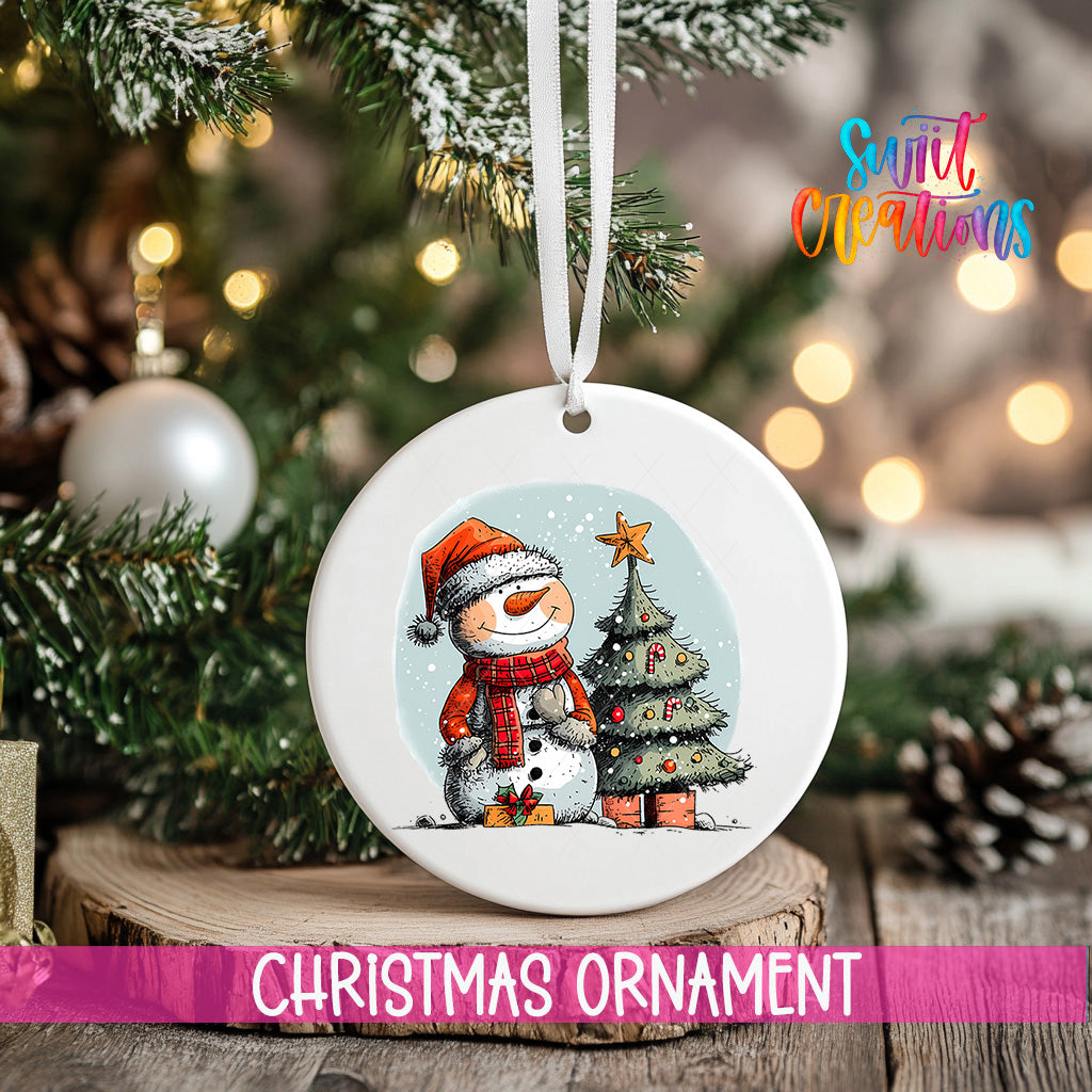 A white Christmas ornament featuring a snowman and a Christmas tree, hanging on a tree branch with a ribbon.