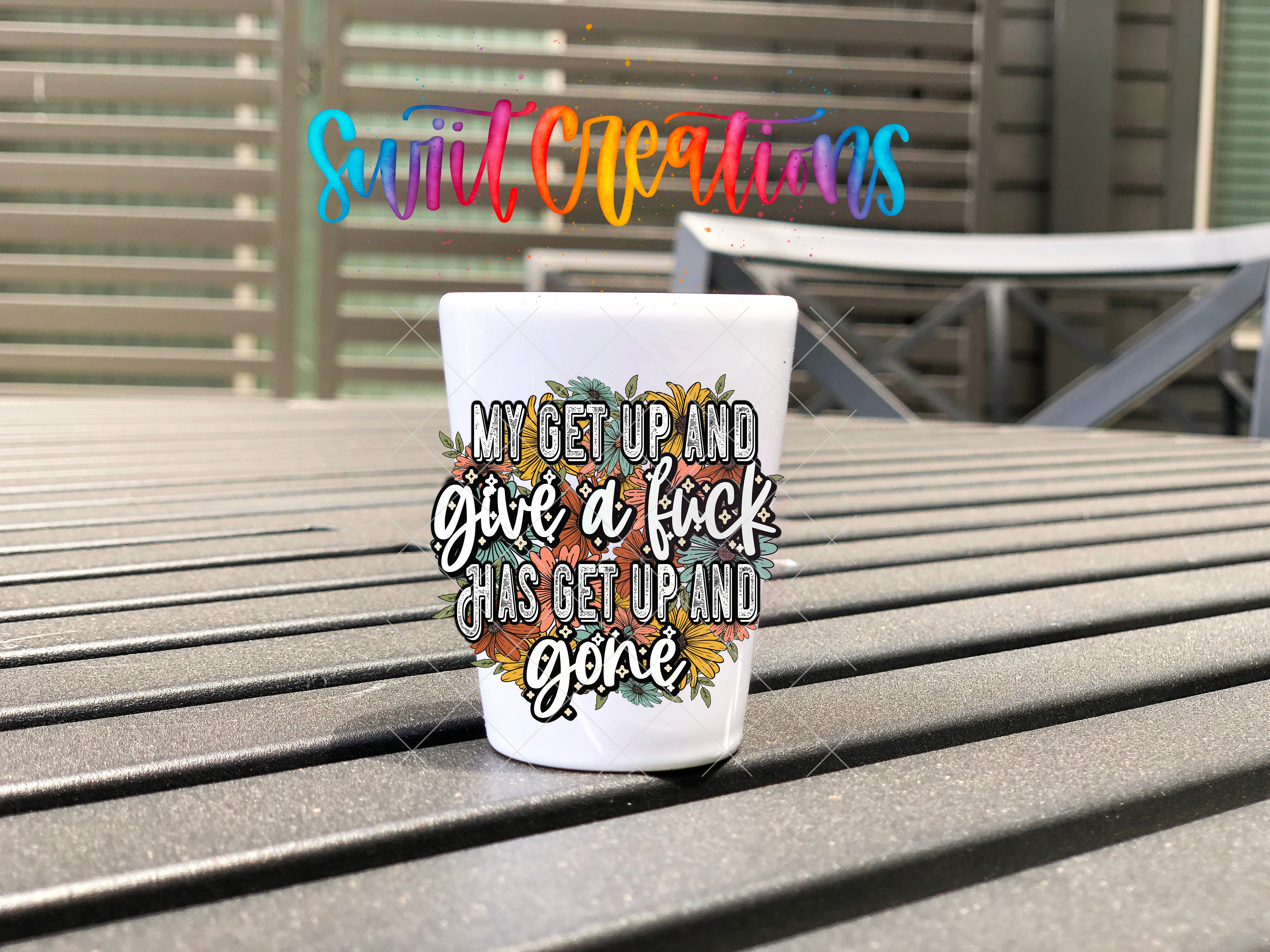 A white shot glass with a colorful, hand-drawn design and text printed on it, placed on a wooden surface.