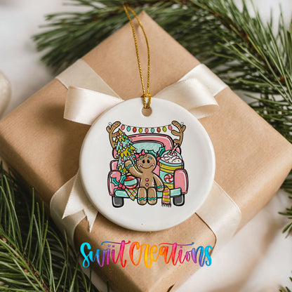 A white ceramic ornament with a gingerbread man and a Christmas tree is hanging from a ribbon on a gift box, surrounded by pine branches.