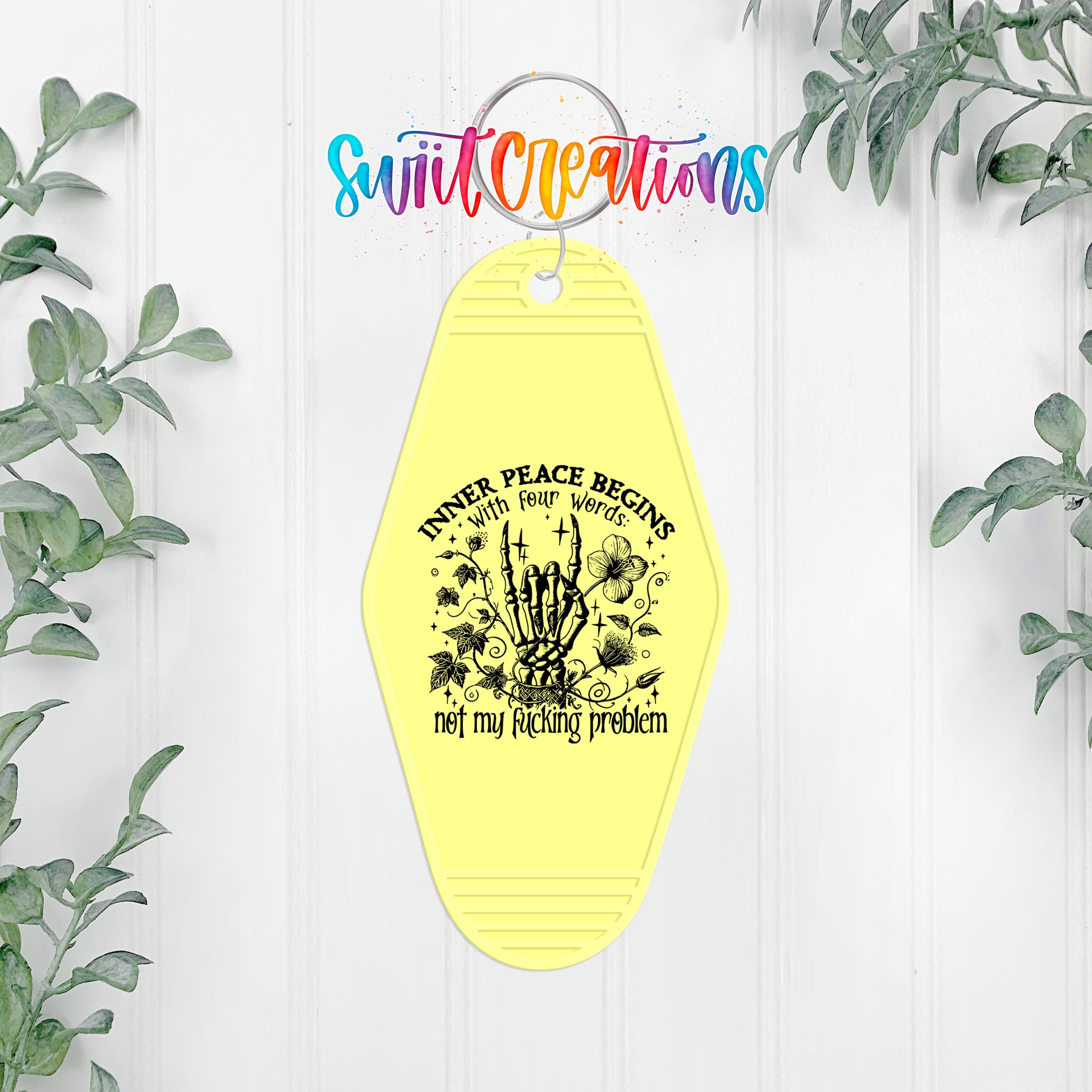 A yellow keychain with a graphic design and text, hanging on a white wall with greenery in the background.