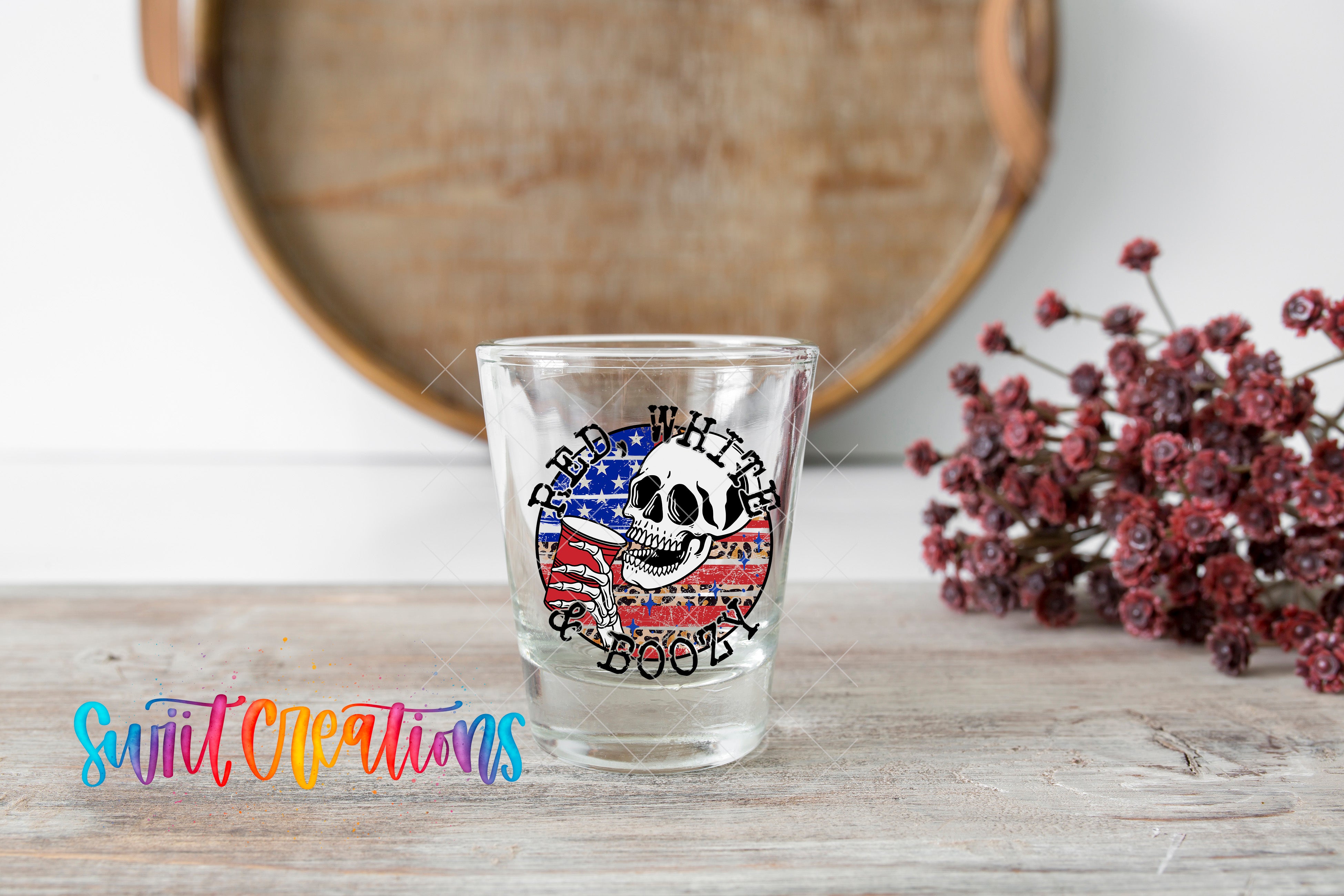 A shot glass with a skull design and an American flag pattern on it, placed on a wooden surface next to a bunch of red flowers.