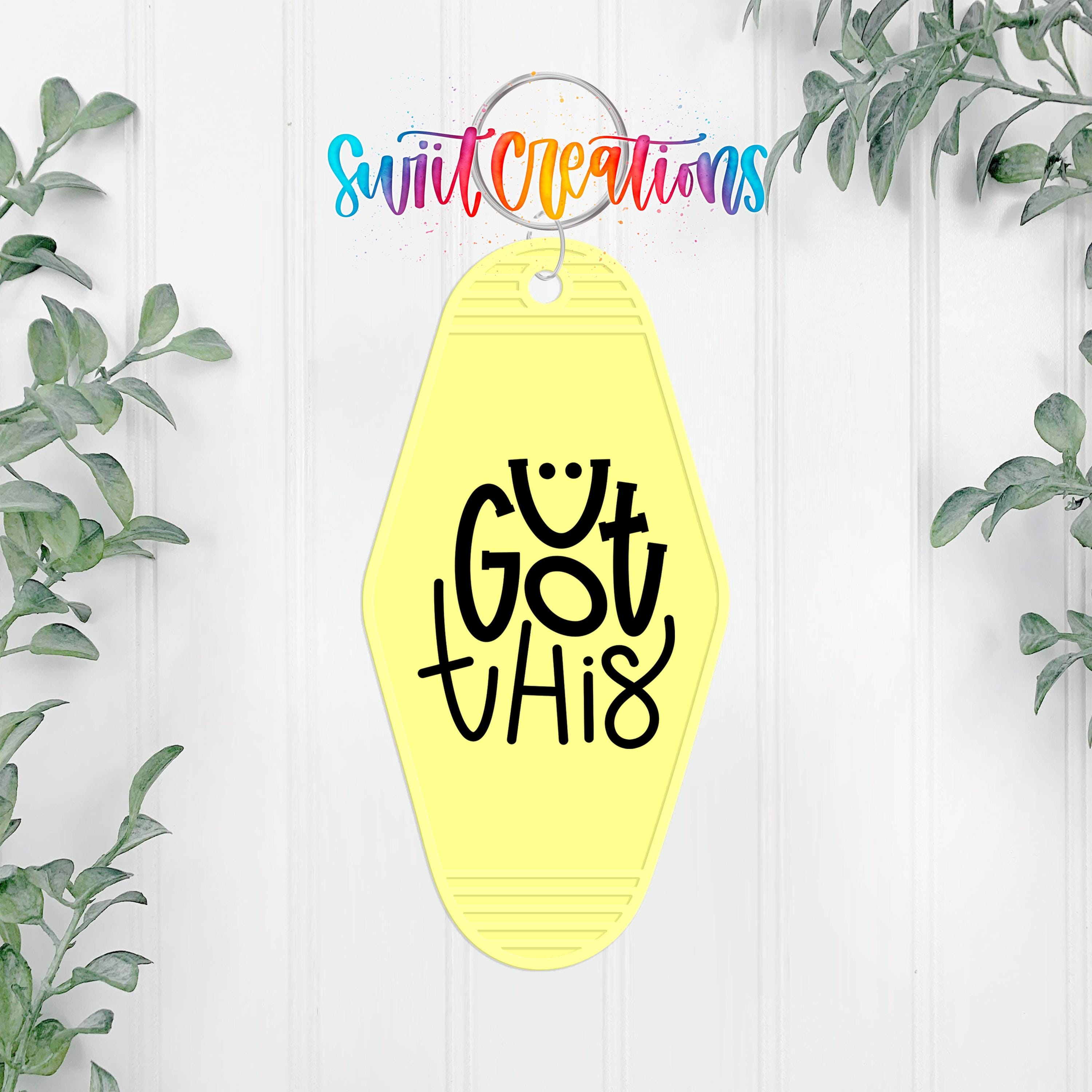A yellow keychain with the text " Got this" printed on it, hanging on a white wall with greenery in the background.