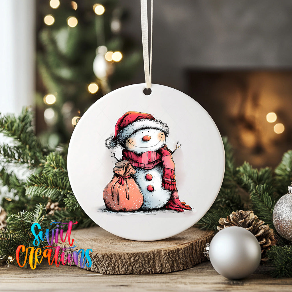 A white ceramic ornament featuring a snowman wearing a red scarf and hat, with a bag around its neck, is displayed on a wooden surface. The ornament is surrounded by Christmas decorations, including a Christmas tree and ornaments.