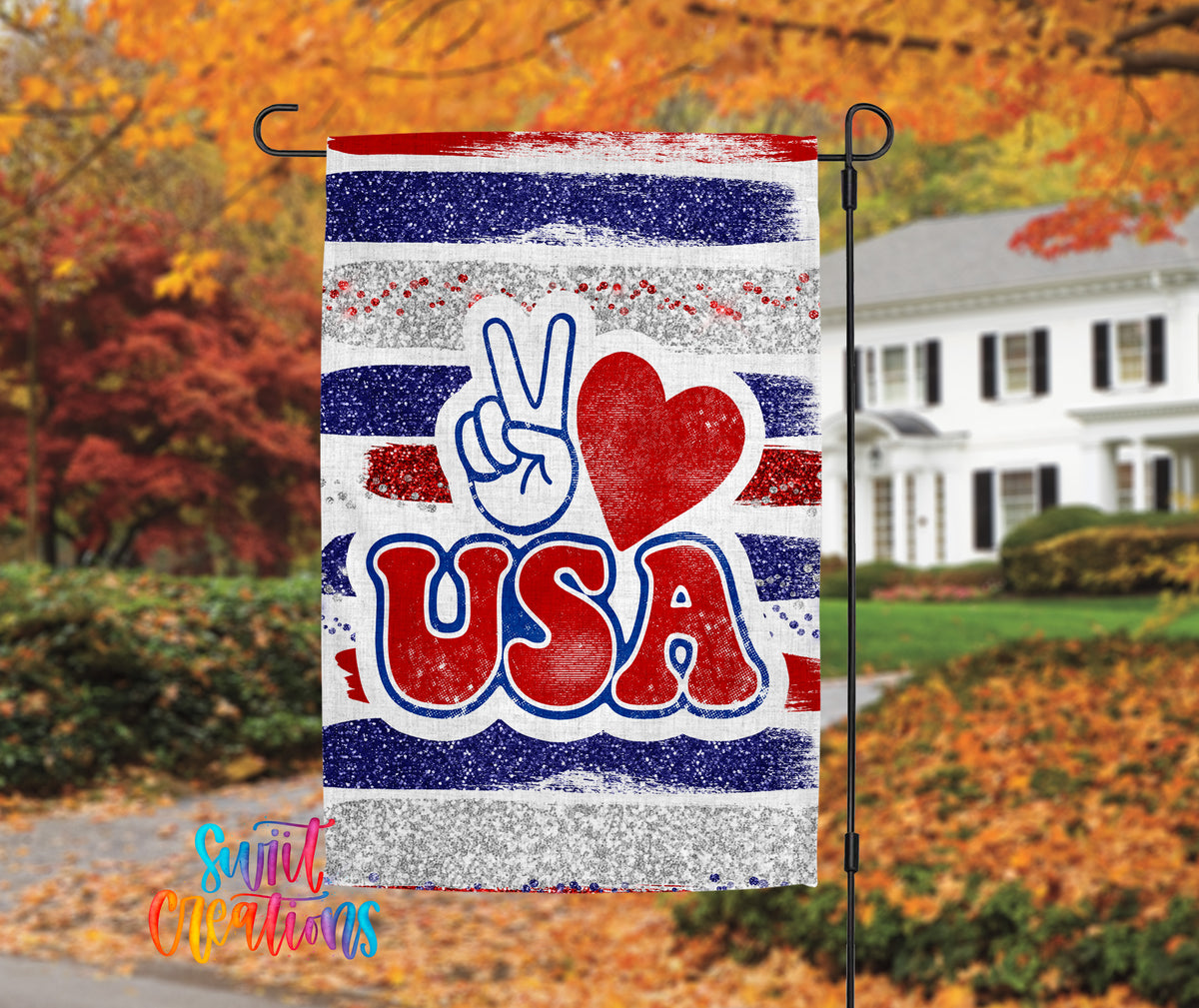 A patriotic flag with the word "USA" prominently displayed, featuring a peace sign and a red heart symbol. The flag is hanging on a black pole, with a house and autumn foliage visible in the background.