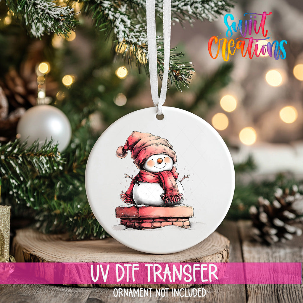 A white ornament with a snowman design hangs on a Christmas tree, surrounded by pine branches and twinkling lights.