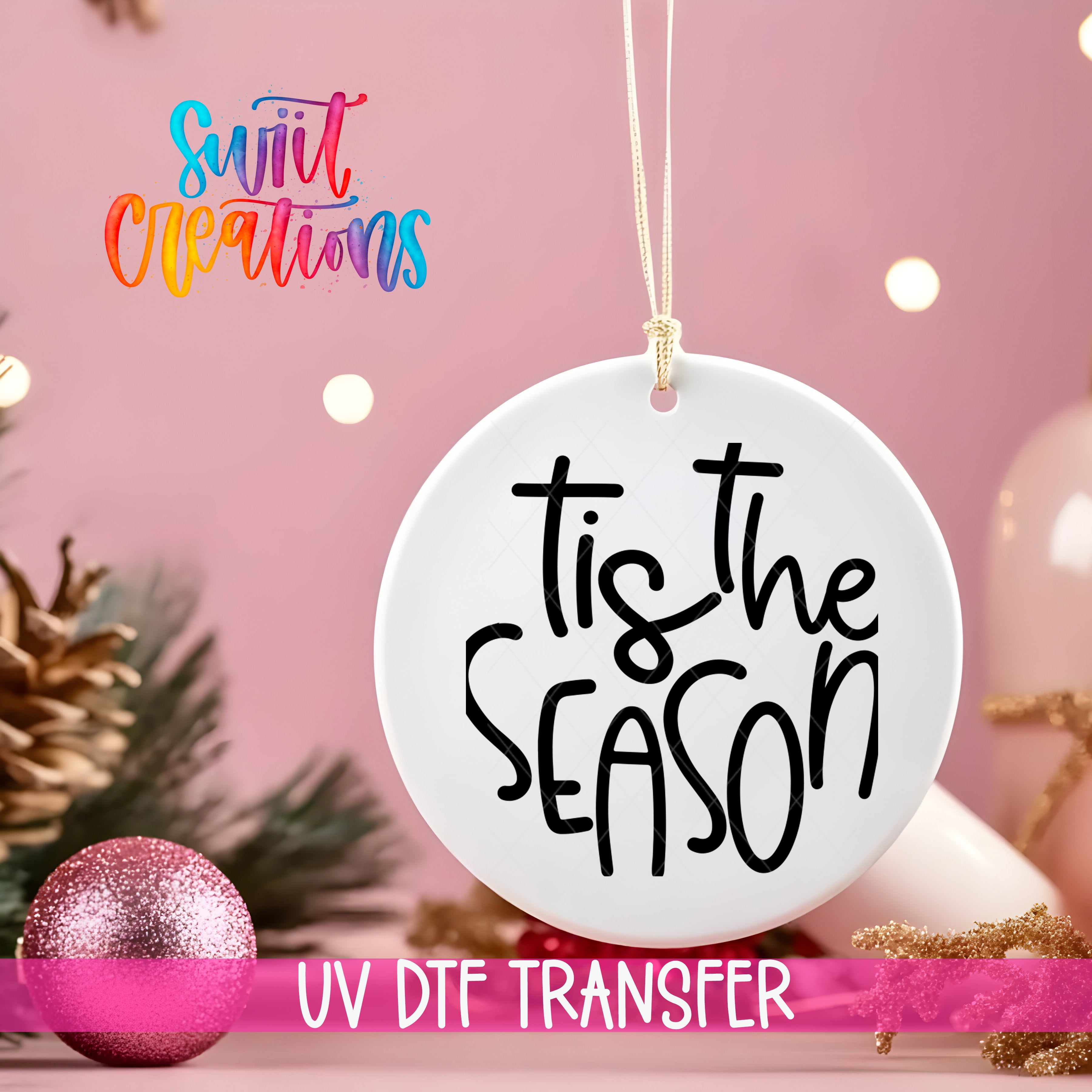 A white ornament with the text "Tis the SEASON" written on it, hanging against a pink background with Christmas decorations and lights in the background.