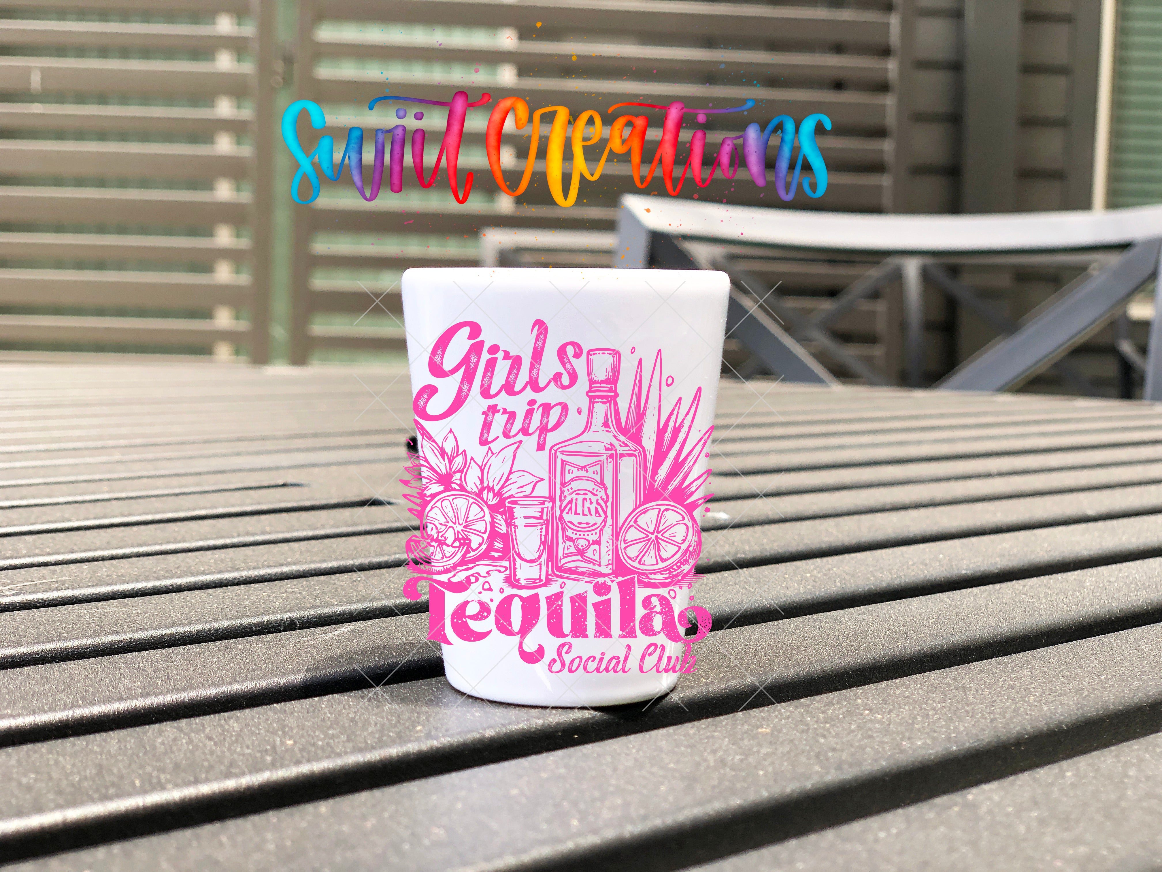 A white shot glass with a pink and purple design featuring tequila-related items, such as a tequila bottle and a tequila glass, sits on a wooden surface.