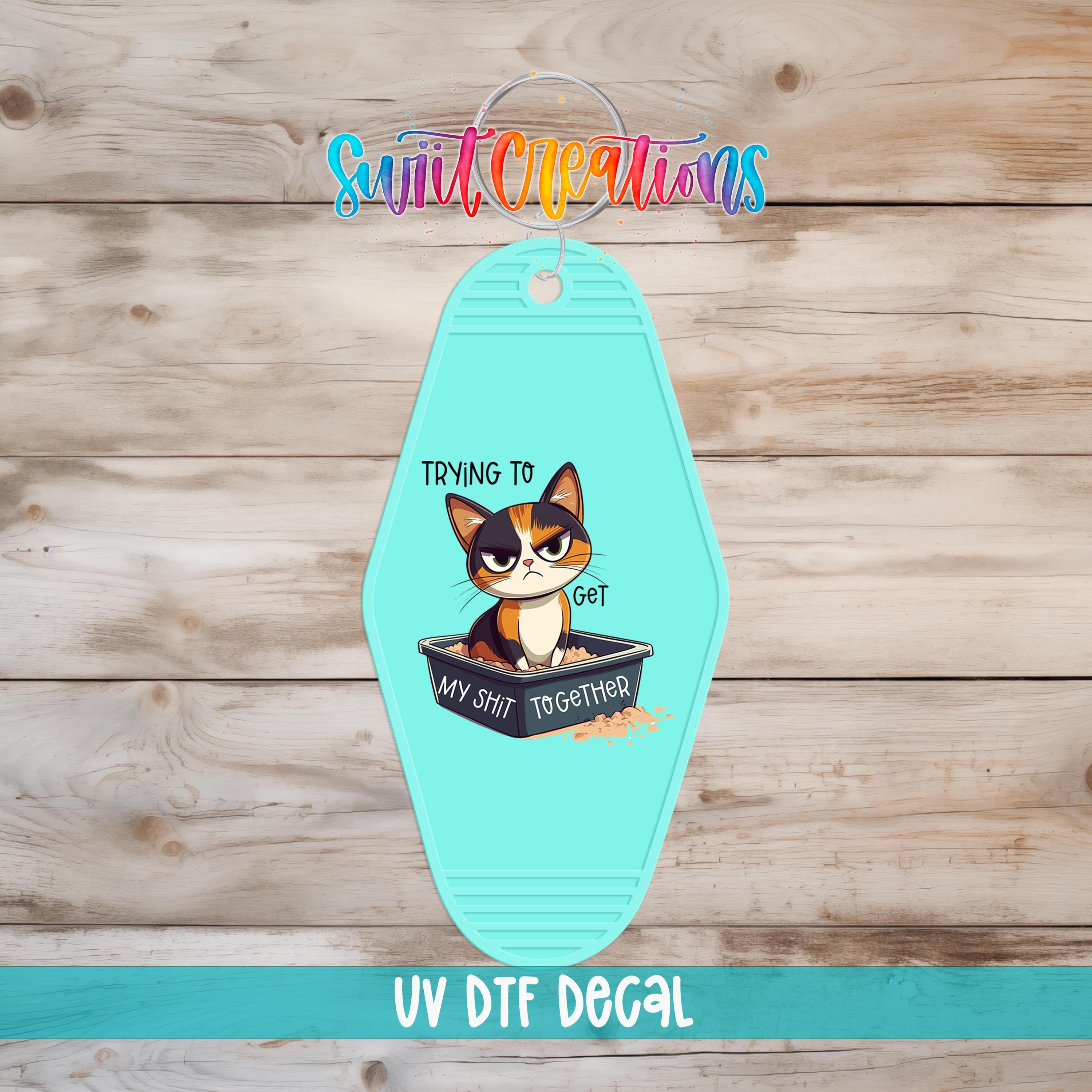 A turquoise keychain with a cartoon cat and the text "Trying to get MY SHIT TOGETHER" printed on it.