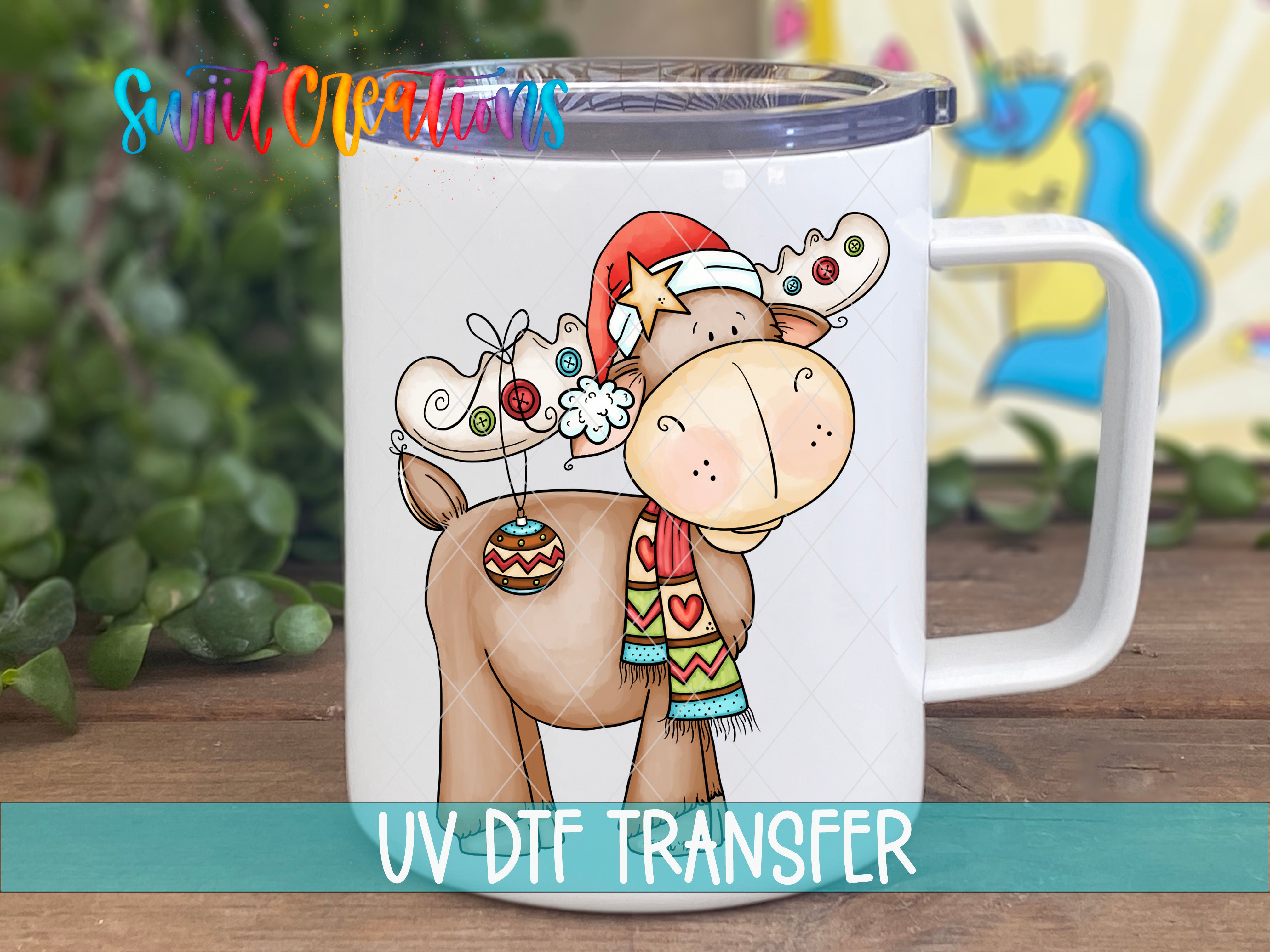 A mug with a cartoon moose wearing a Santa hat and scarf, along with a snowflake design, is displayed on a wooden surface.