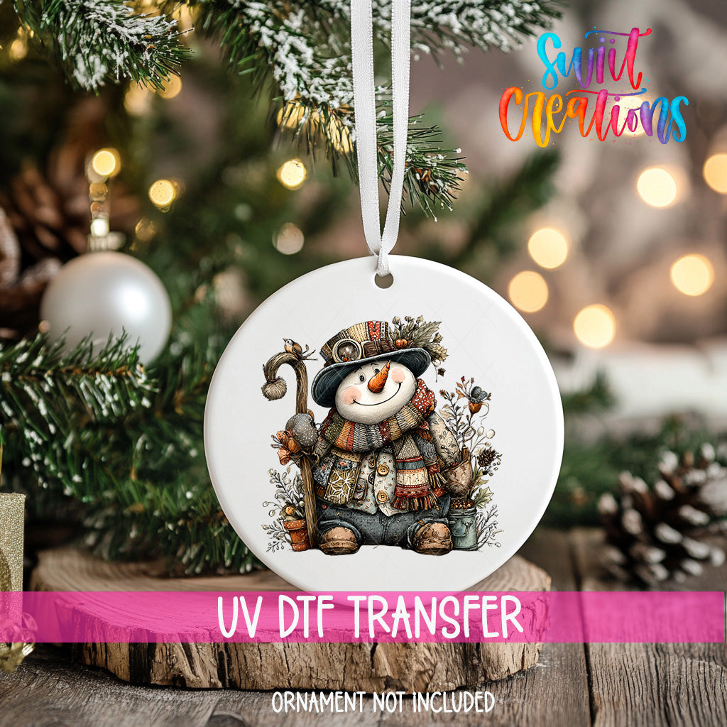A white ornament with a snowman design hangs on a Christmas tree, surrounded by pine branches and other holiday decorations.