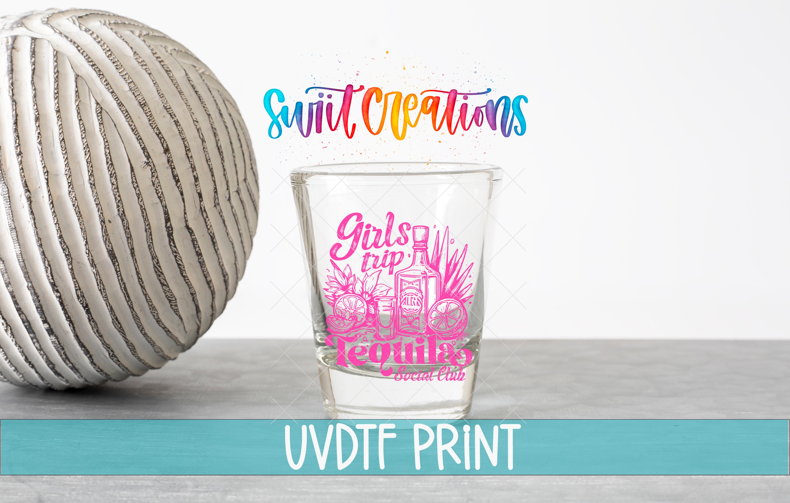 a shot glass with a pink design and text, placed next to a white ball on a gray surface.