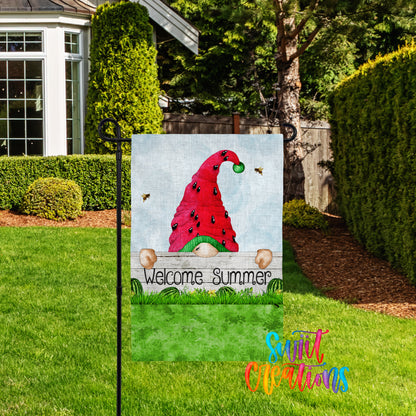 A garden sign featuring a red gnome hat with a green brim and the text "Welcome Summer" is displayed in a grassy yard with a house and trees in the background.