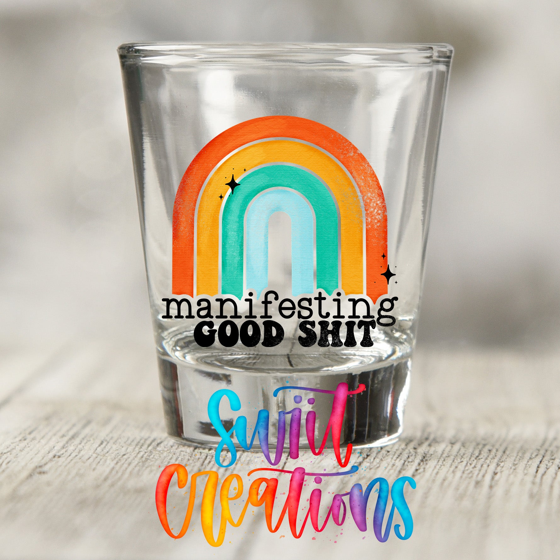 A shot glass with a colorful rainbow graphic and the text "manifesting good shit" printed on it.