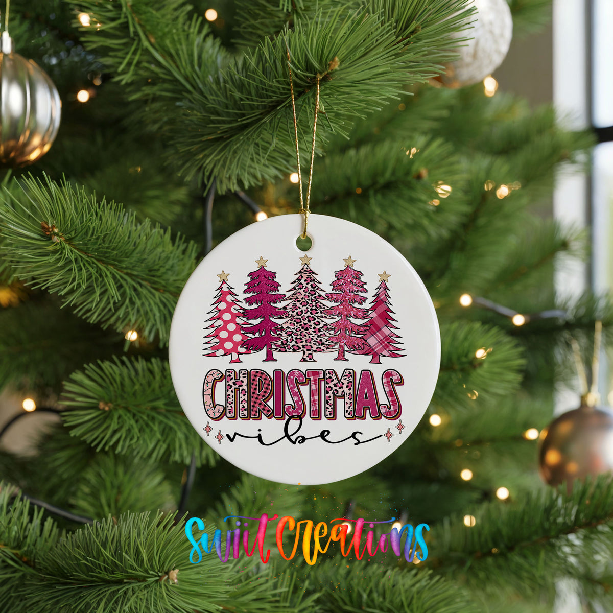A white Christmas ornament with a pink and leopard print design hangs on a pine tree, surrounded by other ornaments and twinkling lights.