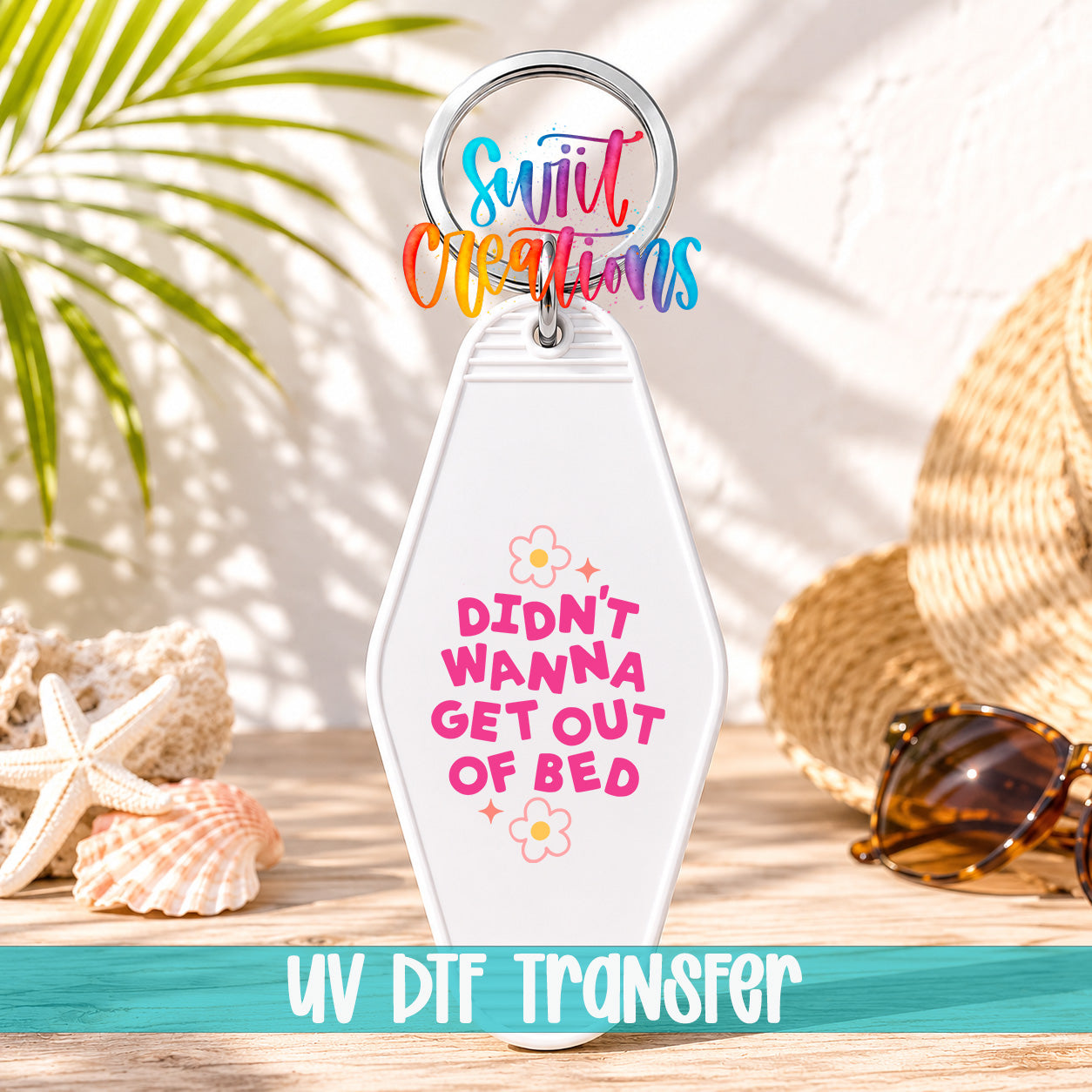 A keychain with a humorous text that says "DIDN'T WANNA GET OUT OF BED" in a playful font, surrounded by sunglasses and a straw hat on a beach-themed background.