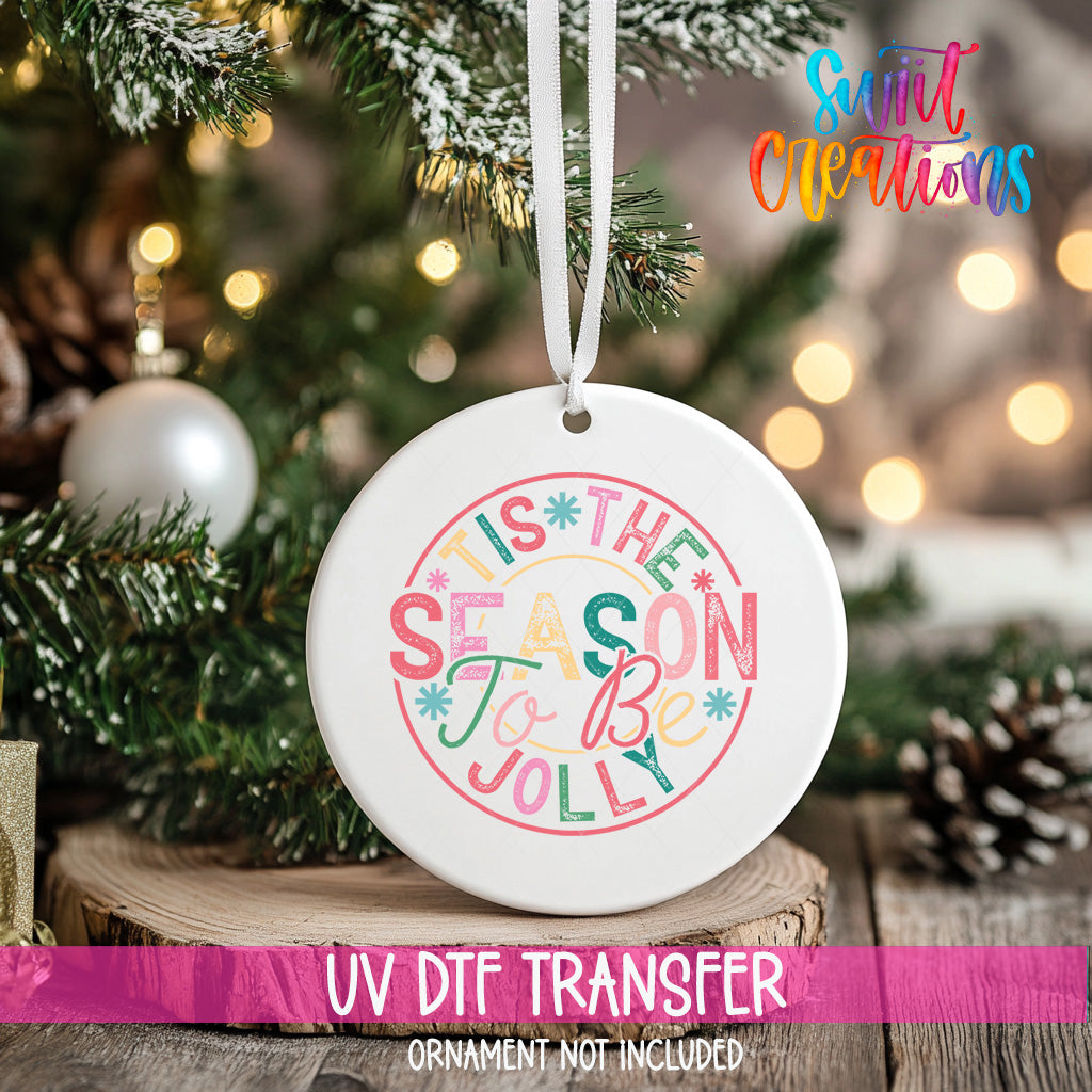 A white ceramic ornament with a colorful design hangs on a Christmas tree, surrounded by pine branches and other holiday decorations.