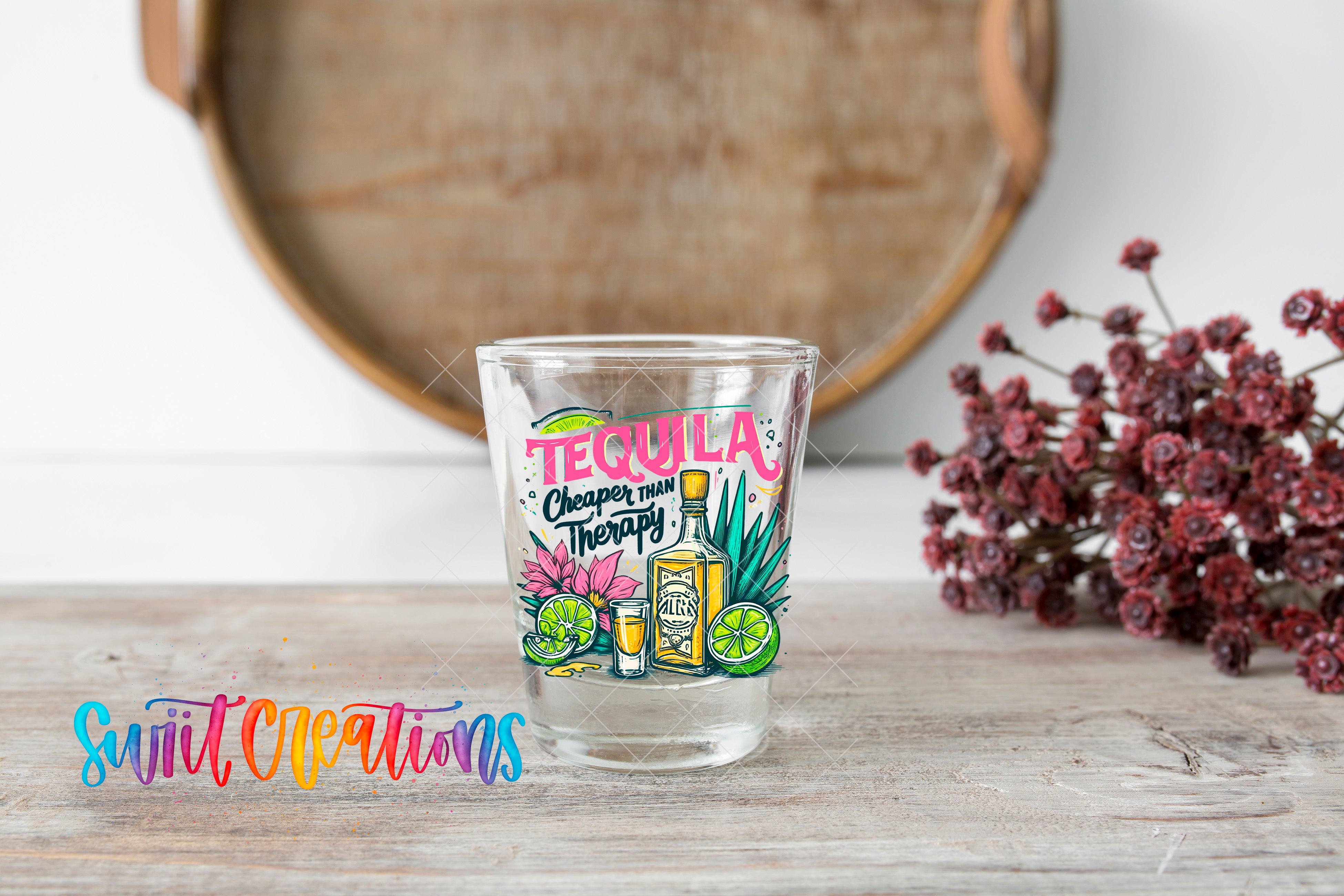A shot glass with a colorful design featuring tequila, lime, and flowers, placed on a wooden surface next to a bunch of red flowers.