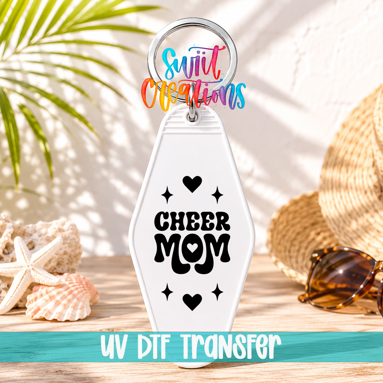 A keychain with a message that says "CHEER MOM" in black text, surrounded by hearts and stars, placed on a wooden surface with sunglasses and a straw hat in the background.
