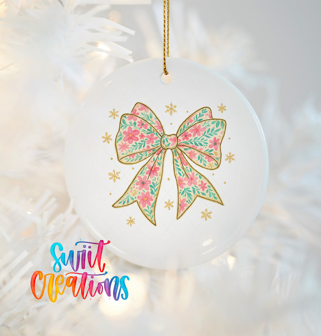 A white ceramic ornament with a floral bow design and gold accents, hanging on a white background with a blurred Christmas tree visible.