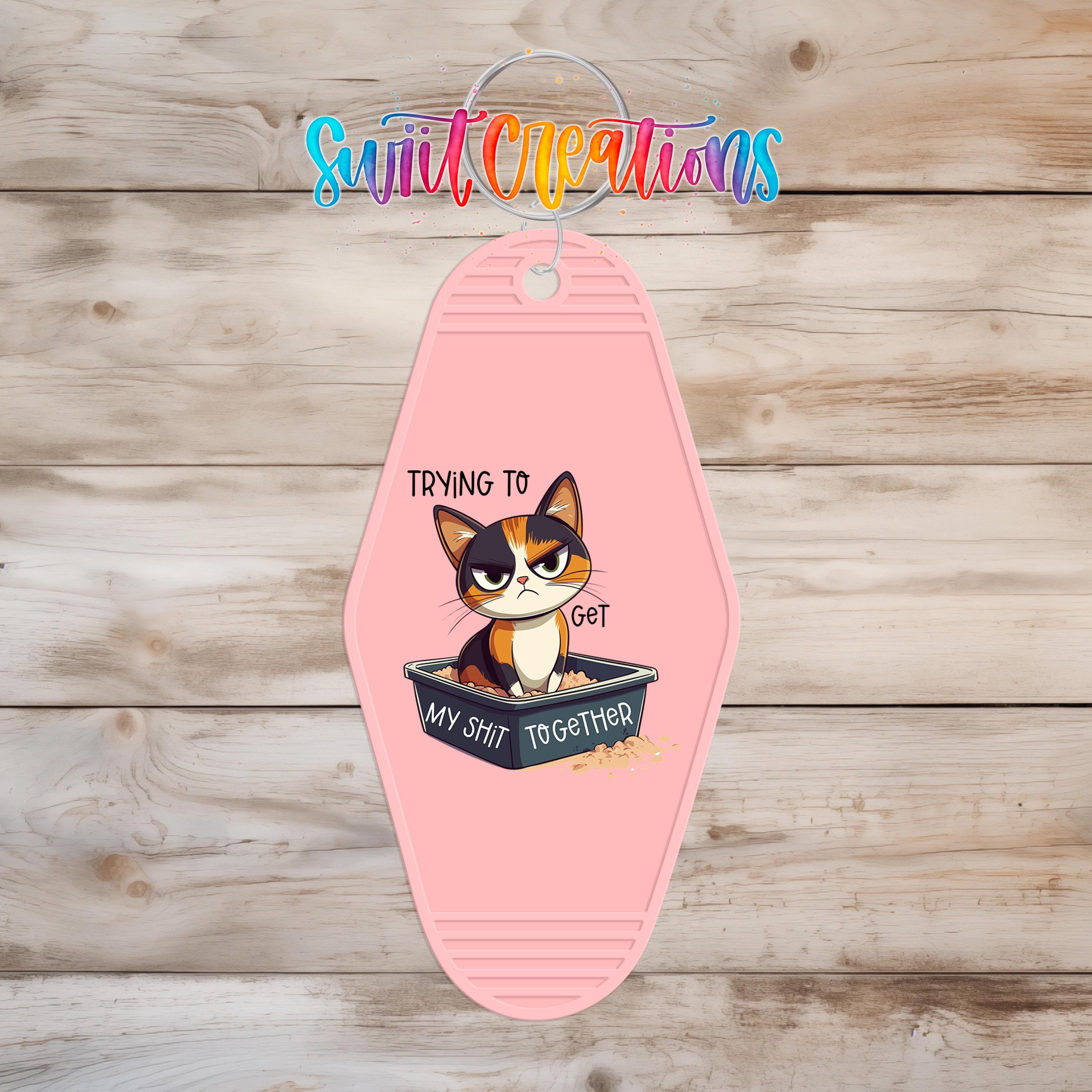 A pink keychain with a cartoon cat and the text "Trying to Get My Shit Together" printed on it.