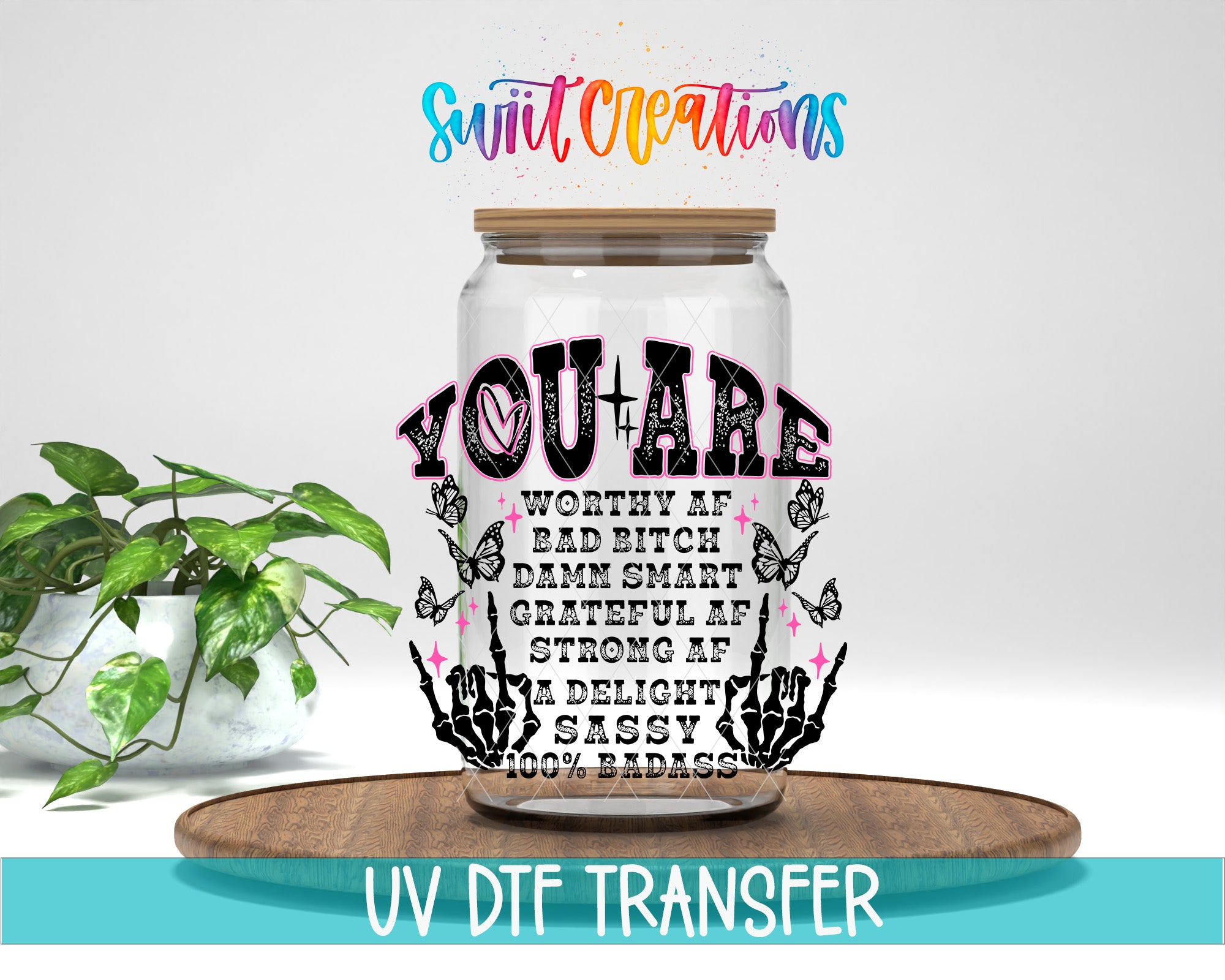 A glass jar with a message inside, placed on a wooden surface. The jar has a lid and is surrounded by a plant and some butterflies. The text on the jar reads "YOU ARE WORTHY OF BAD BITCH DRAMA SMART GRATEFUL OF STRONG AF DELIGHT SASSY 100% BADASS".